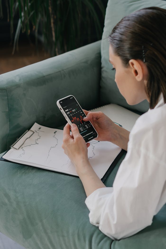 A woman examines financial stock data on her smartphone, sitting indoors with a graph on paper.